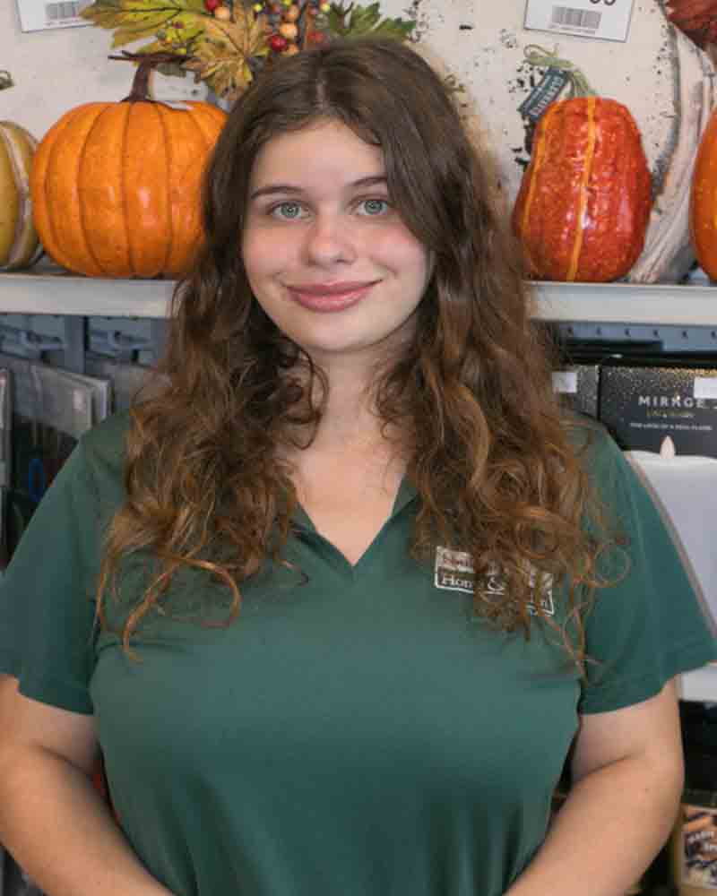 A young woman with long, wavy hair stands in front of autumn decorations.