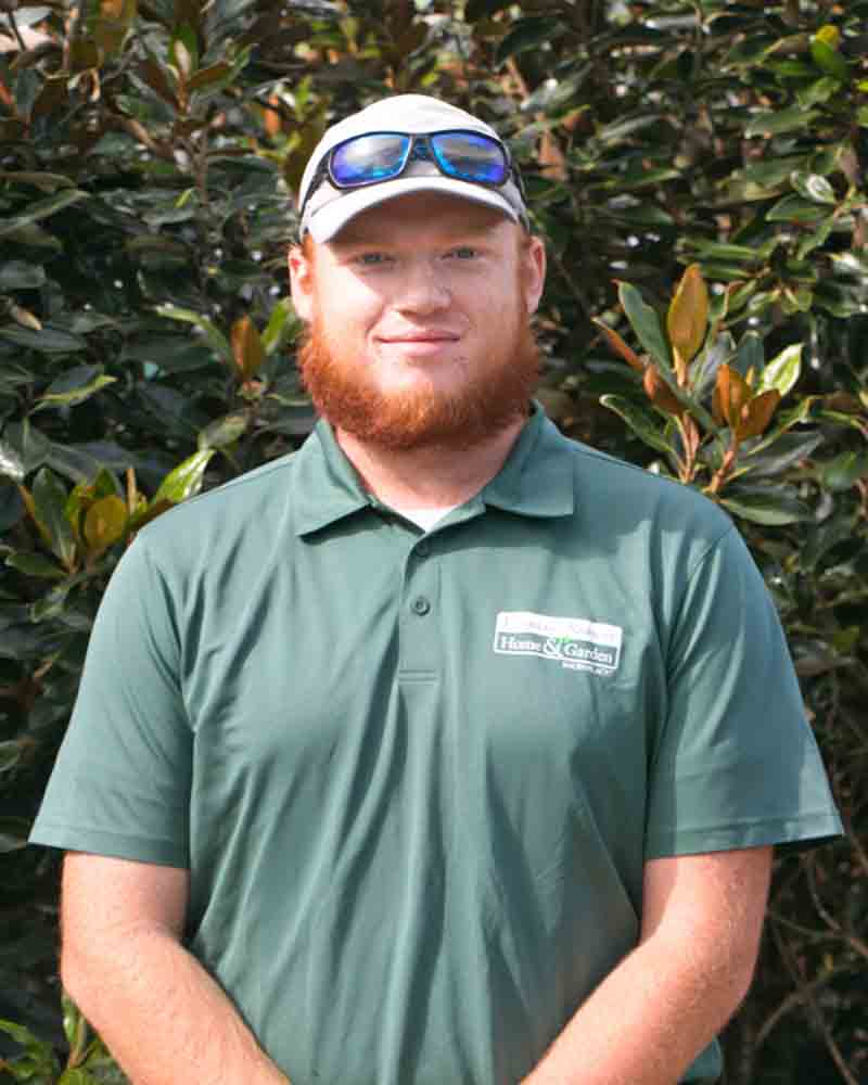 A man with a red beard stands in front of green foliage, wearing a green shirt and cap.