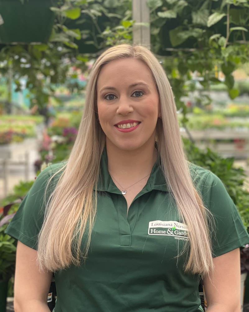 A woman with long blonde hair smiles, wearing a green polo shirt in a greenhouse.