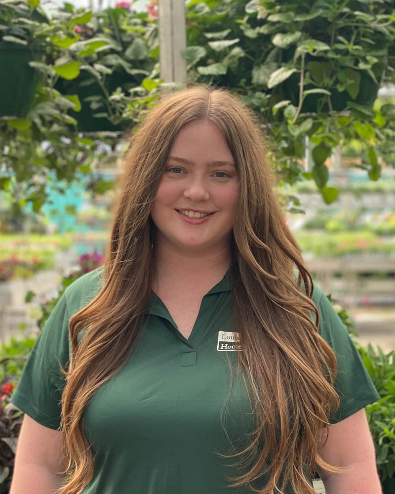 A smiling woman with long hair stands in a greenhouse surrounded by plants.