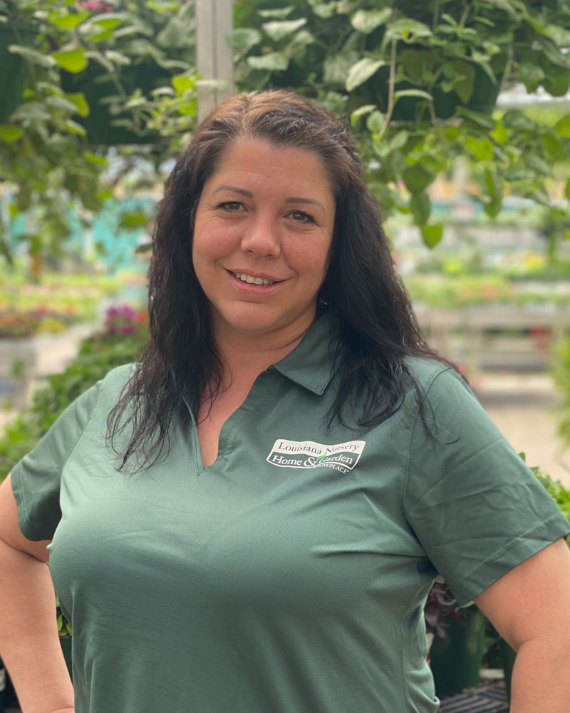 A woman with long dark hair smiles, wearing a green polo shirt in a garden setting.