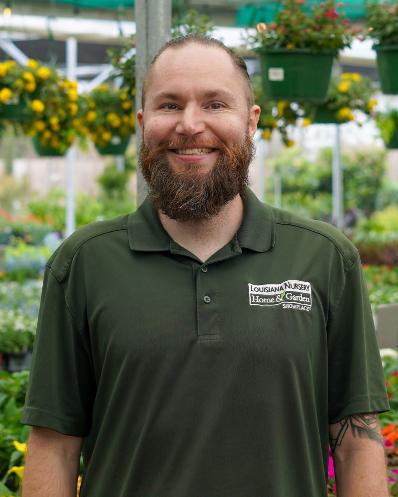 Smiling man with a beard in a green shirt, surrounded by plants and flowers.