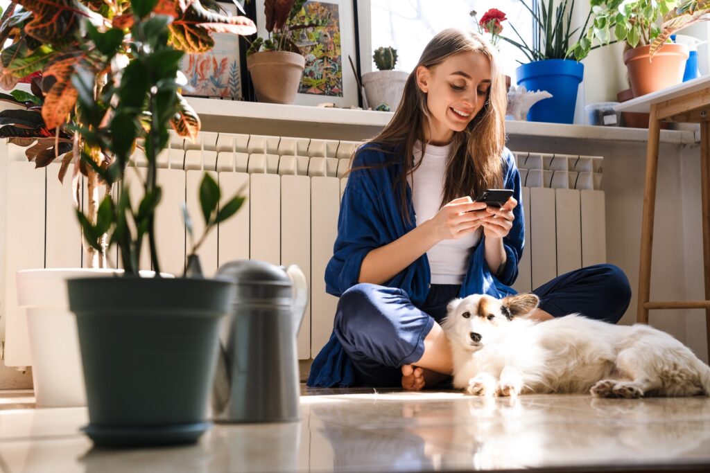 Woman with her dog, using a smartphone amid house plants Baton Rouge loves, basking in sunlight on the floor of a cozy room.